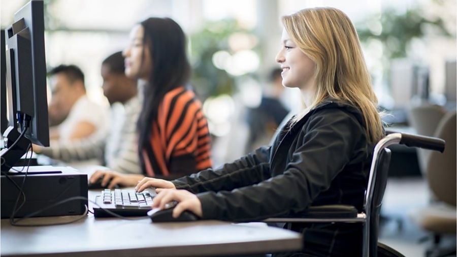 En una mesa con varias computadoras y usuarios, una chica con discapacidad sonríe mientras utiliza la computadora 