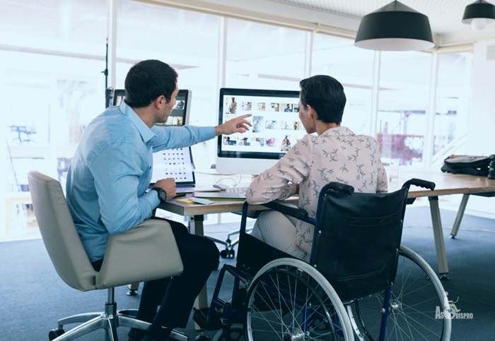 Un joven en silla de ruedas es instruido por una persona sin discapacidad frente a un escritorio y una computadora. 