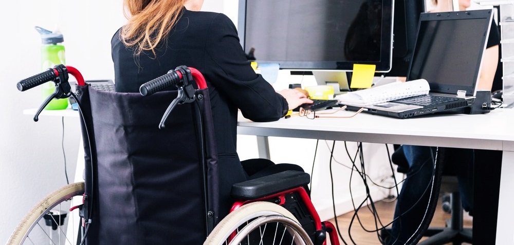 Una mujer en silla de ruedas está de espaldas trabajando frente a una computadora en su escritorio.