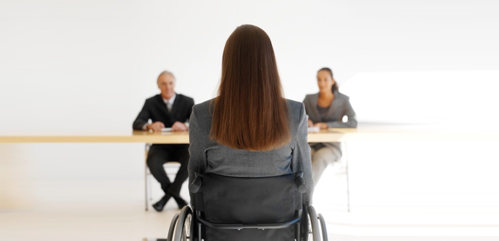 Una mujer de pelo largo, sentada en su silla de ruedas, esta de frente a 2 reclutadores en una entrevista de trabajo.